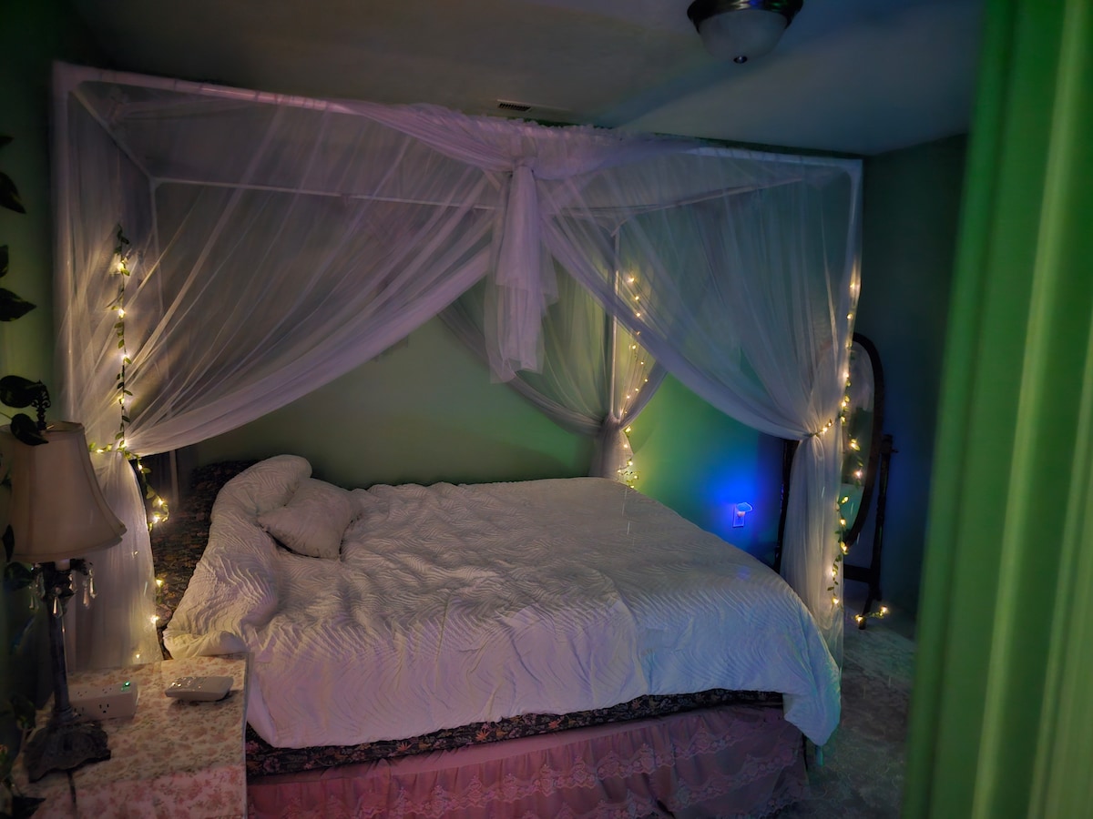 A softly lit bedroom features a canopy bed adorned with sheer white curtains and string lights, creating a serene ambiance. The walls are painted in a gentle green, and a full-length mirror is positioned nearby. A bedside lamp contributes to the cozy atmosphere.