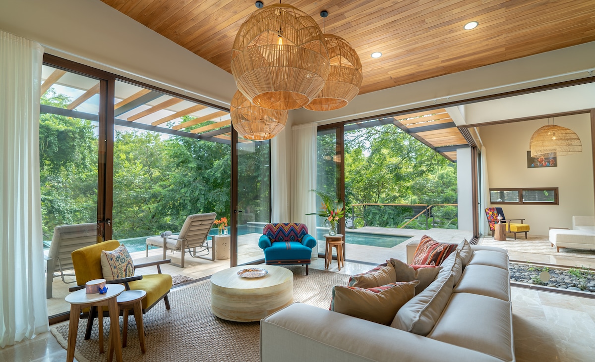 A spacious living area features large glass doors that open to lush greenery and a sparkling pool. Comfortable seating includes a colorful chair and a sofa adorned with soft pillows. Natural wood accents and warm light from woven pendant lamps enhance the inviting atmosphere.