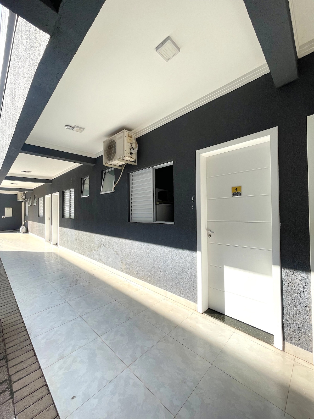 A corridor is displayed, featuring a series of individual doors painted in white, set against a dark gray wall. Windows with horizontal slats allow natural light to filter in. An air conditioning unit is mounted on the wall, contributing to a comfortable environment.