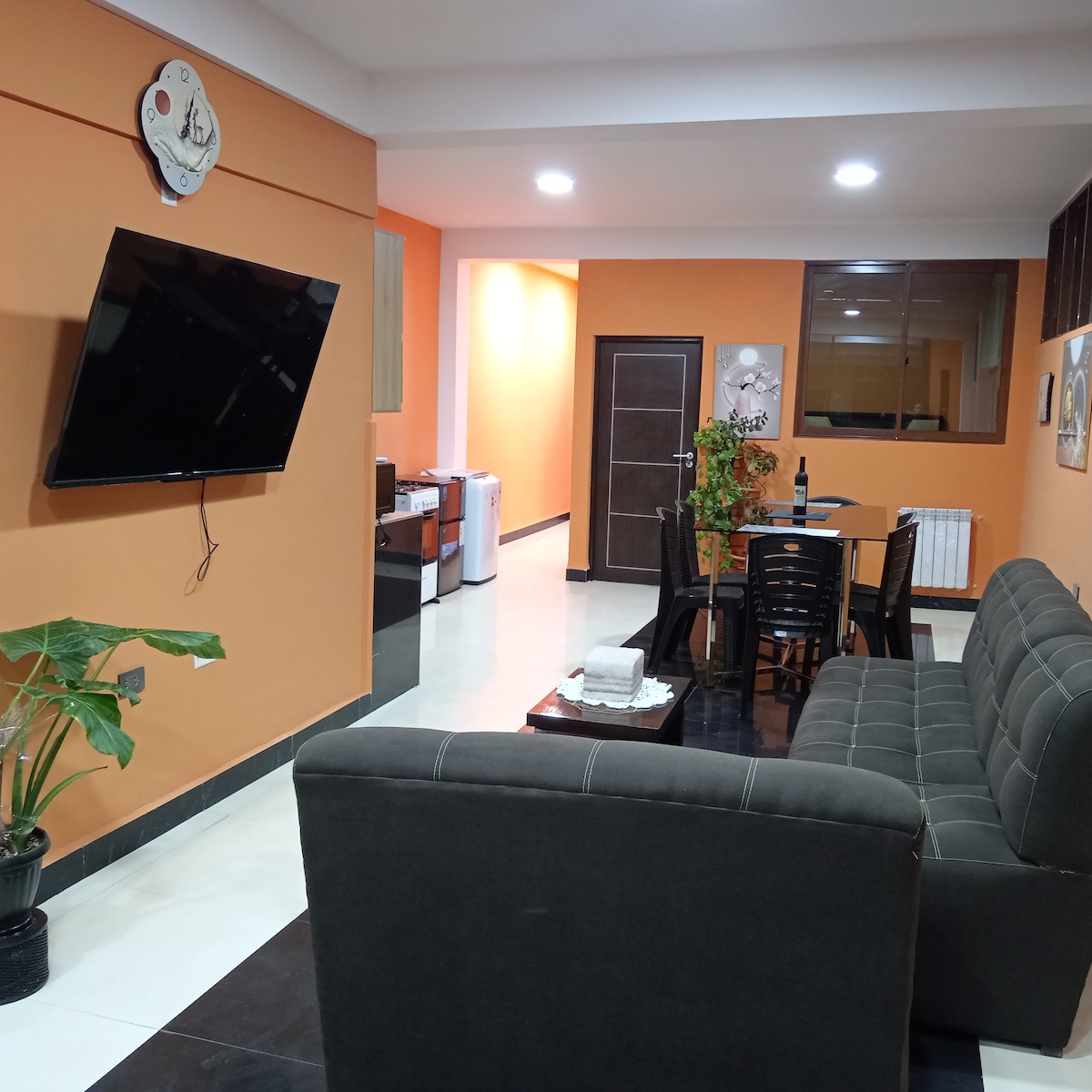 A living area features an earthy brown sofa and a wall-mounted television. The space is brightened by orange walls and includes a dining table with several chairs. A small indoor plant stands at the side, adding a touch of greenery to the room.