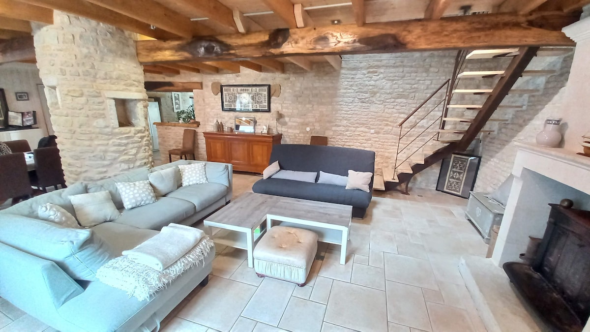 A spacious living area features a comfortable sectional sofa and a sleek coffee table. Stone walls and wooden beams contribute to the rustic charm. A staircase with a modern railing leads to the upper level, while a cozy fireplace adds warmth to the space.
