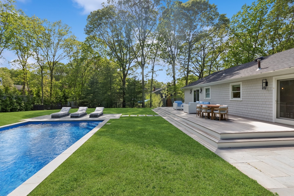 A spacious backyard features a heated pool surrounded by a stone deck. Lounge chairs are placed nearby on the well-manicured lawn. A dining area with a table and chairs sits adjacent to the house, offering a view of the swimming pool and the lush greenery beyond.