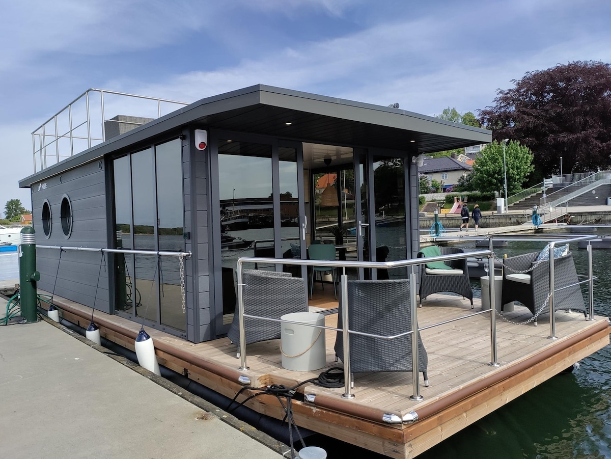 A modern houseboat is docked at a marina, featuring large windows that offer views of the surroundings. An outdoor seating area with wicker chairs is positioned on the deck, inviting relaxation. The boat's sleek design is complemented by a spacious upper deck.
