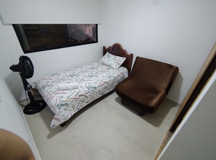 Habitación secundaria con cama sencilla, silla tipo puff y ventilador. Espacio cómodo y funcional para el descanso de un huésped adicional.