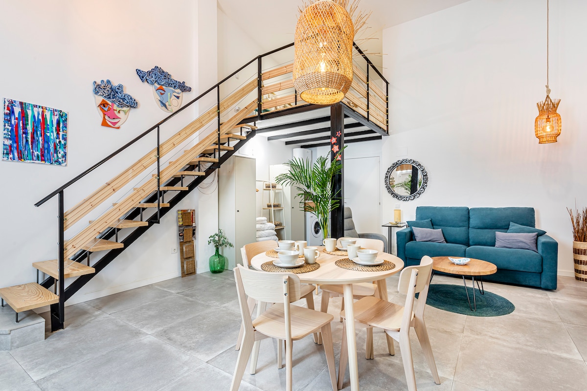 An open living area features a modern dining table set for four, complemented by a spacious seating area with a teal sofa. A decorative circular rug adds warmth, while a staircase leads to an upper level. Colorful artwork and plants enhance the inviting environment.