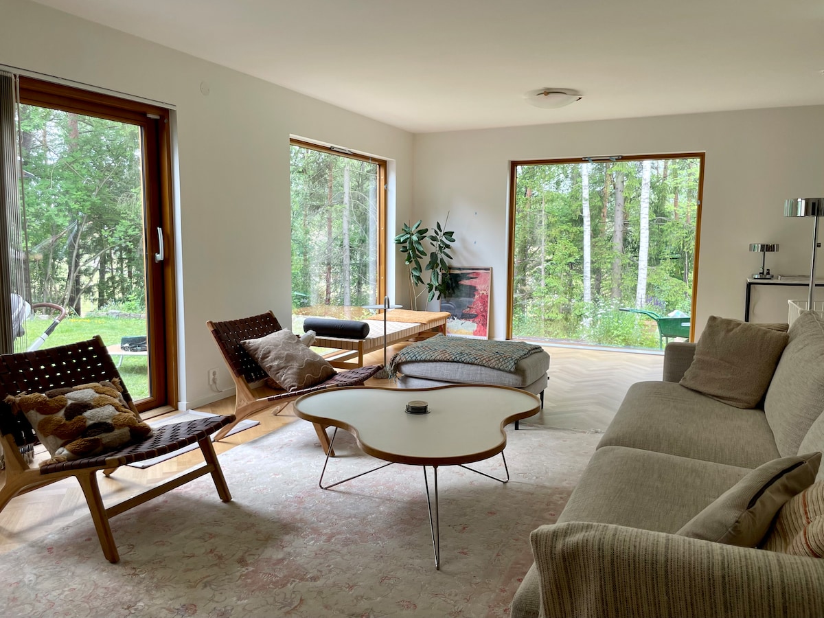 A comfortable living area is highlighted by large windows overlooking a forested landscape. The space features a sectional sofa, a round coffee table, and a mix of seating options, including a lounge chair and cushions. Natural light fills the room, creating a serene atmosphere.