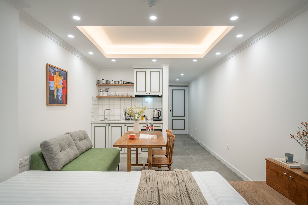 A modern studio apartment showcases a spacious living area with a green sofa and wooden dining table set for two. The kitchen features white cabinetry and essential cookware arranged neatly. Natural light illuminates the room, highlighting the contemporary décor and a colorful wall art piece.