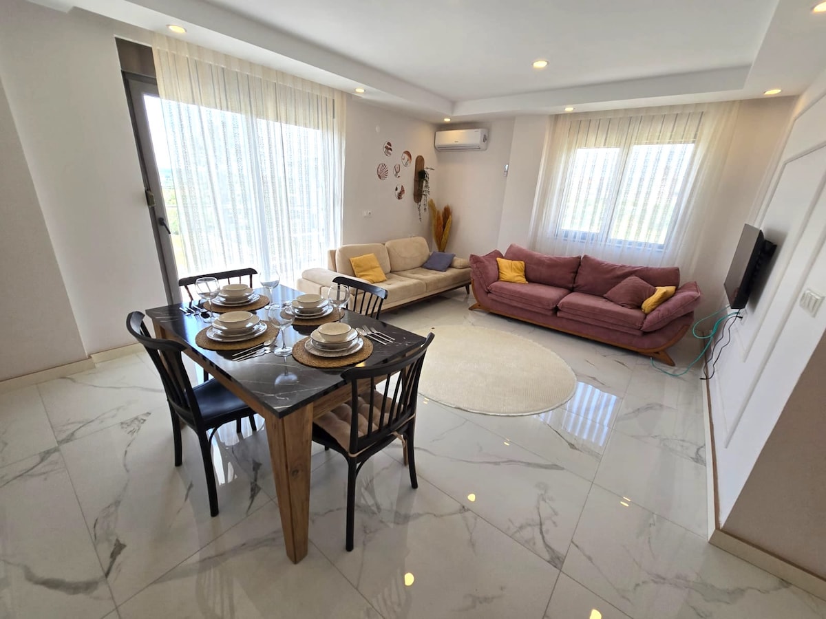 A modern living space features a wooden dining table set for four, complemented by a plush seating area with two sofas in neutral tones. Natural light fills the room through large windows, while elegant curtains provide a cozy ambiance.