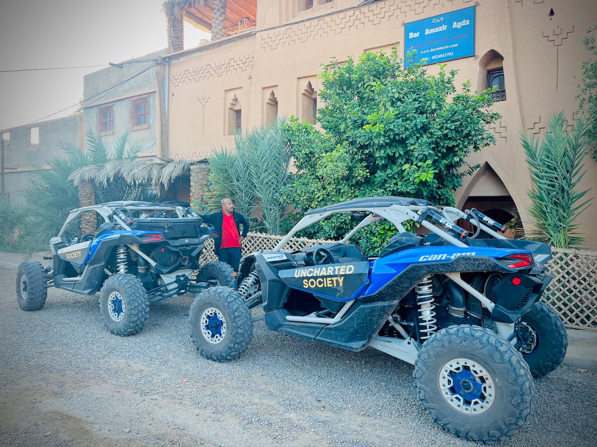 Two off-road vehicles with distinctive markings are parked alongside a gravel path. The backdrop features traditional Moroccan architecture, complemented by lush palm trees and local flora. A warm, inviting ambiance is created by the guesthouse's rustic exterior.
