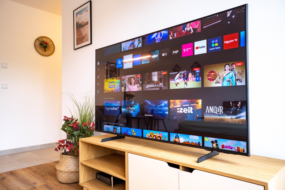 A large smart TV is displayed on a wooden console, showcasing various streaming service options. Decorative plants in a woven basket are positioned nearby. The bright space is enhanced by neutral walls and minimalistic decor.