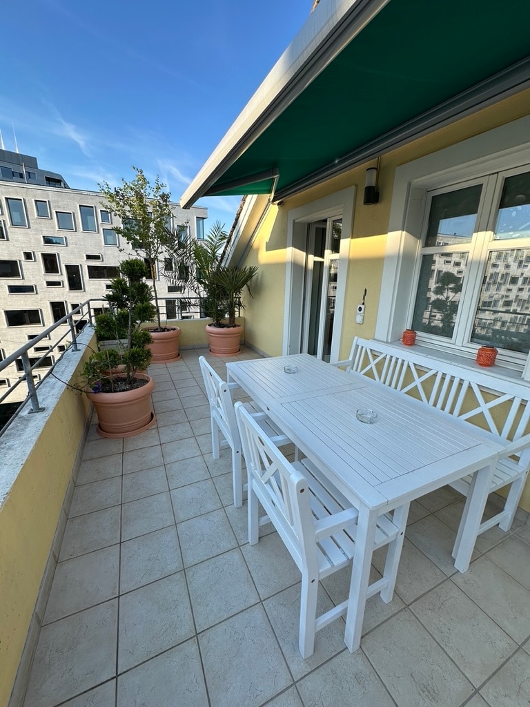 A balcony space features a white dining table surrounded by matching chairs, complemented by potted plants. The area offers a view of nearby buildings and is partially shaded by a green awning, with sunny skies visible above.