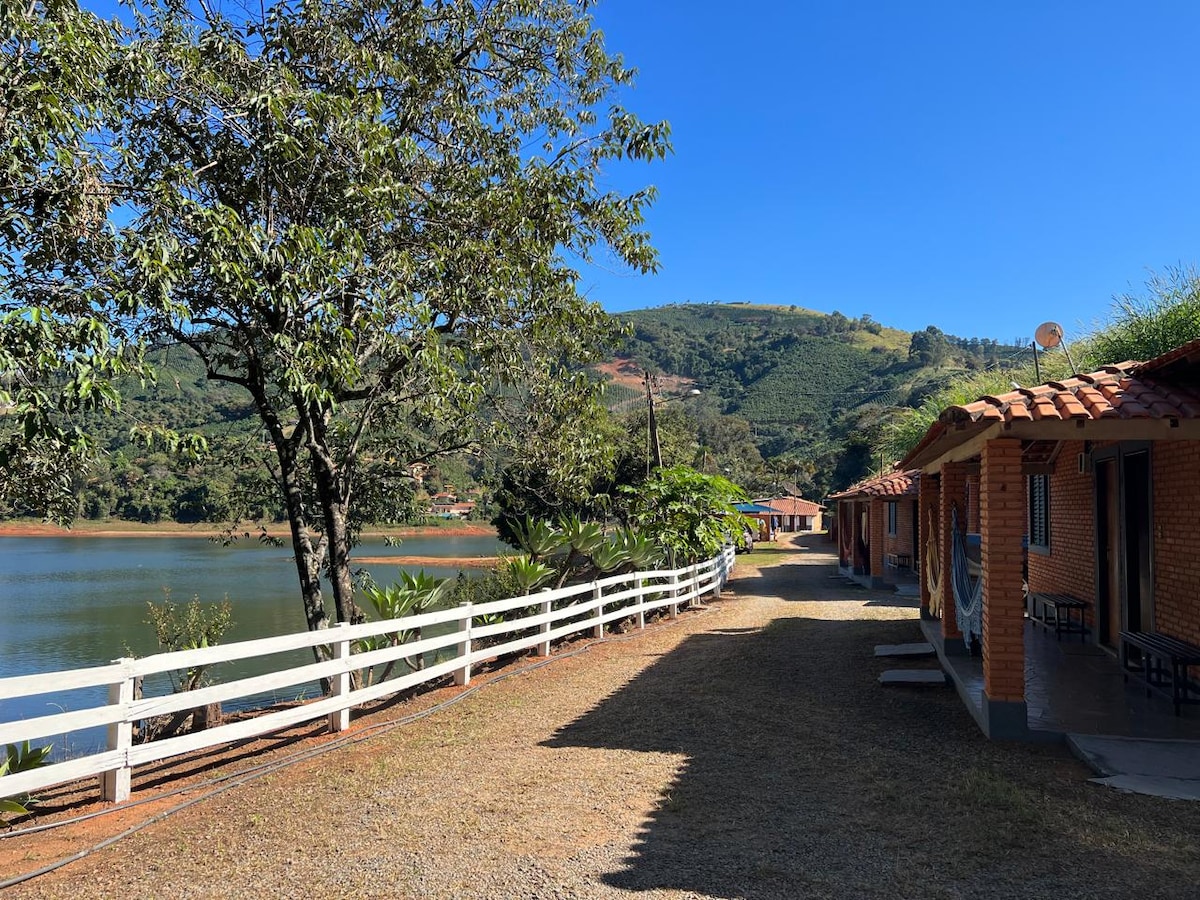A pathway lined with white fencing runs beside a tranquil lake, surrounded by lush greenery and rolling hills. Clustered cabins with red tile roofs can be seen along the path, offering inviting proximity to nature and the serene water.