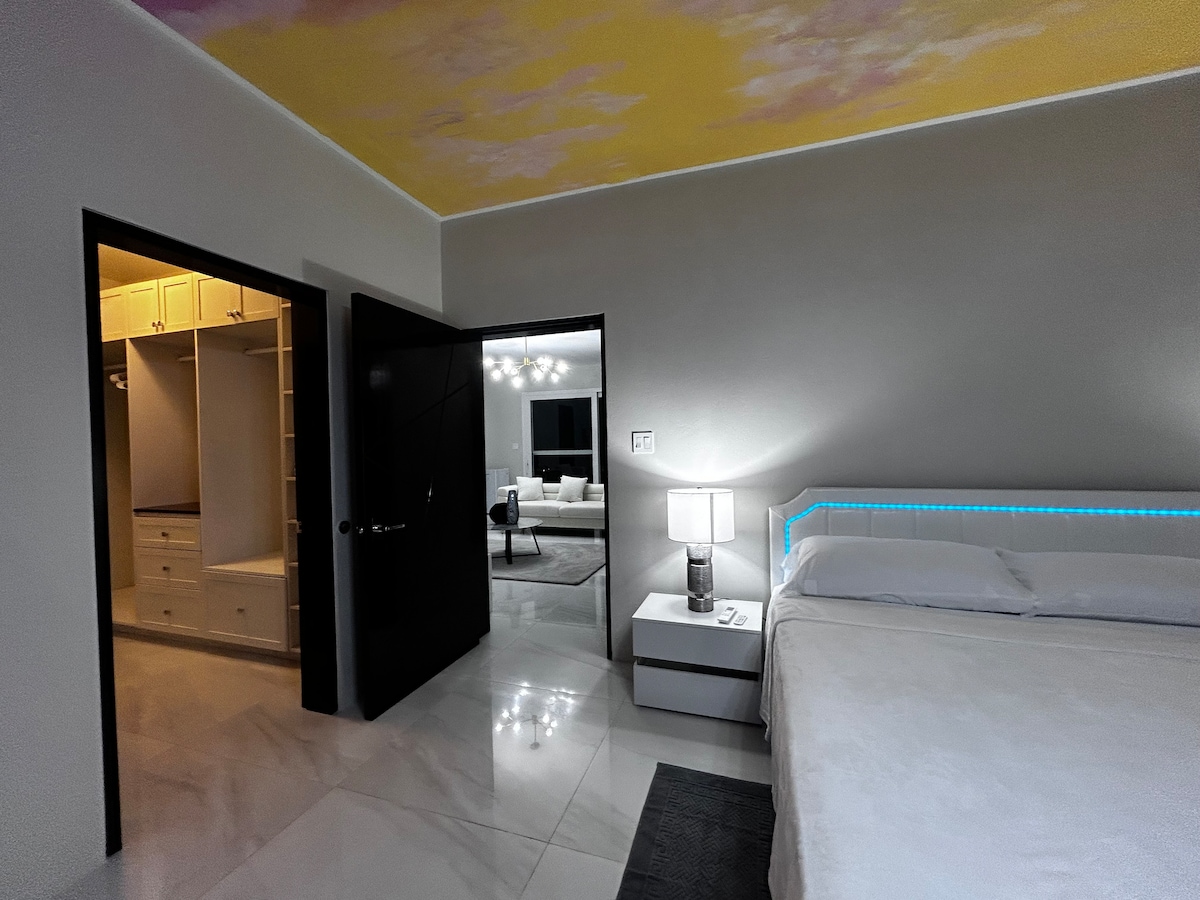A spacious bedroom features a modern bed with illuminated accents on the headboard. A bedside table with a lamp offers soft lighting. A doorway leads to a walk-in closet, and a view into the living area can be seen through an open door.