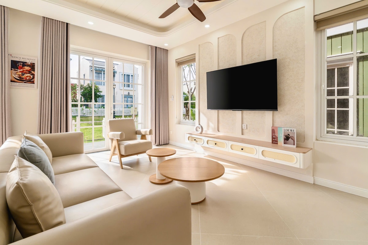 A bright living room features a spacious beige sofa paired with a light wooden coffee table. A large flat-screen television is mounted on the wall opposite the seating area, with natural light streaming in through multiple windows dressed with soft curtains.