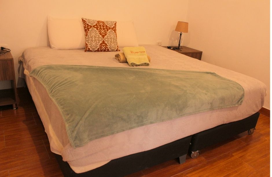 A spacious bed is dressed in neutral linens, featuring decorative pillows for added style. A soft, green throw blanket is draped casually across the bed. Two nightstands with lamps are flanking the bed, and warm wooden flooring adds to the overall ambiance.