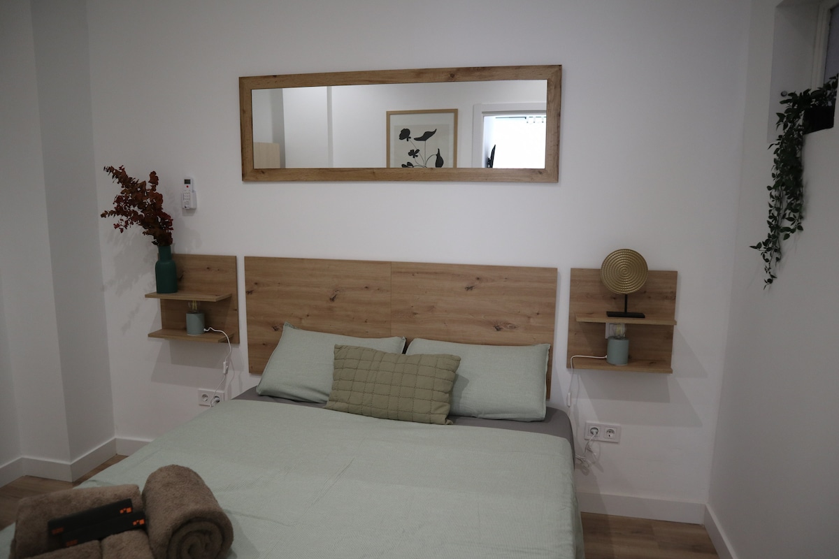 A modern bedroom features a wooden headboard with matching bedside shelves on either side. A mirror hangs above the bed, reflecting the room's light. Soft green bedding complements the neutral tones, while decorative items add subtle accents, enhancing the overall comfort of the space.