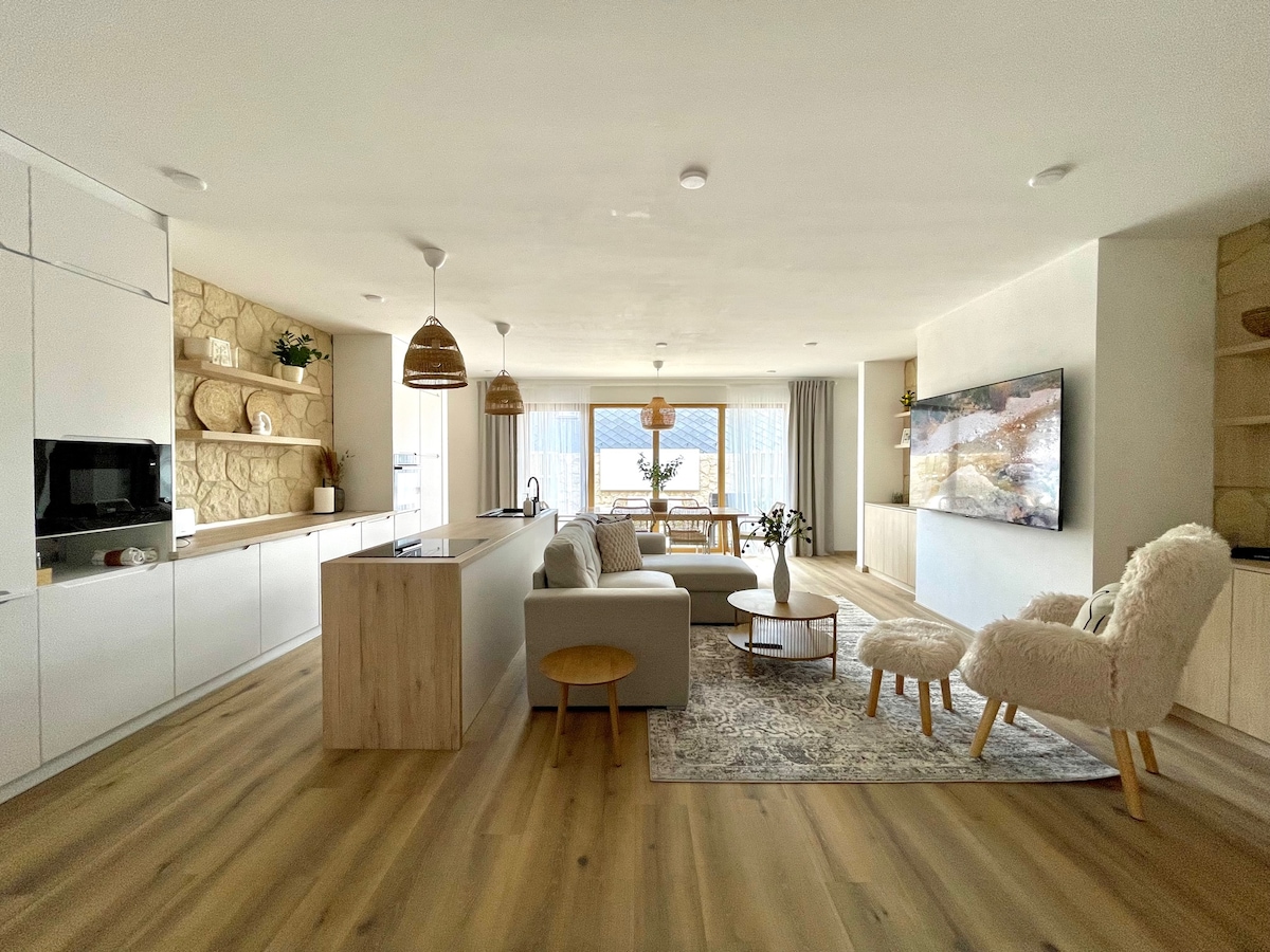 An open-concept living area features a modern kitchen with white cabinetry and wood accents. A spacious sofa and a fluffy chair provide seating around a round coffee table on a patterned rug. Large windows bring in ample natural light, enhancing the inviting space.