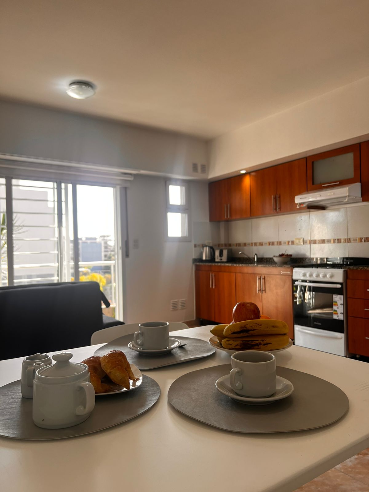 The well-lit kitchen area is equipped with wooden cabinetry and modern appliances. A dining table is set with white dishware, including cups and a serving dish with fruits. Large windows allow natural light to fill the space, enhancing the welcoming atmosphere.