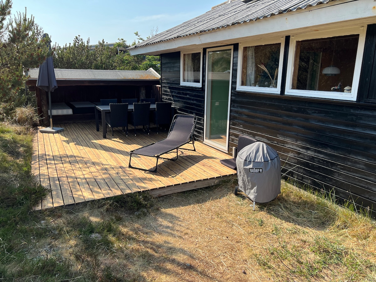 A sunlit terrace extends from the house, featuring outdoor seating and a lounge chair. A sheltered area provides additional seating. The building displays a classic design with large windows, allowing natural light to illuminate the surrounding landscape.