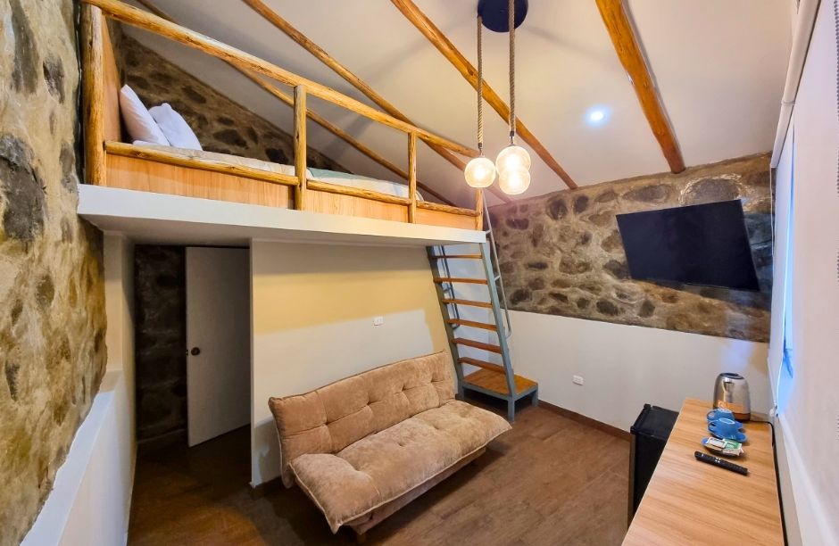 A two-level room is presented with exposed wooden beams and stone walls. A cozy seating area features a light brown sofa. A staircase leads to a lofted sleeping area, which is positioned above the main space. A flat-screen television is mounted on the wall.