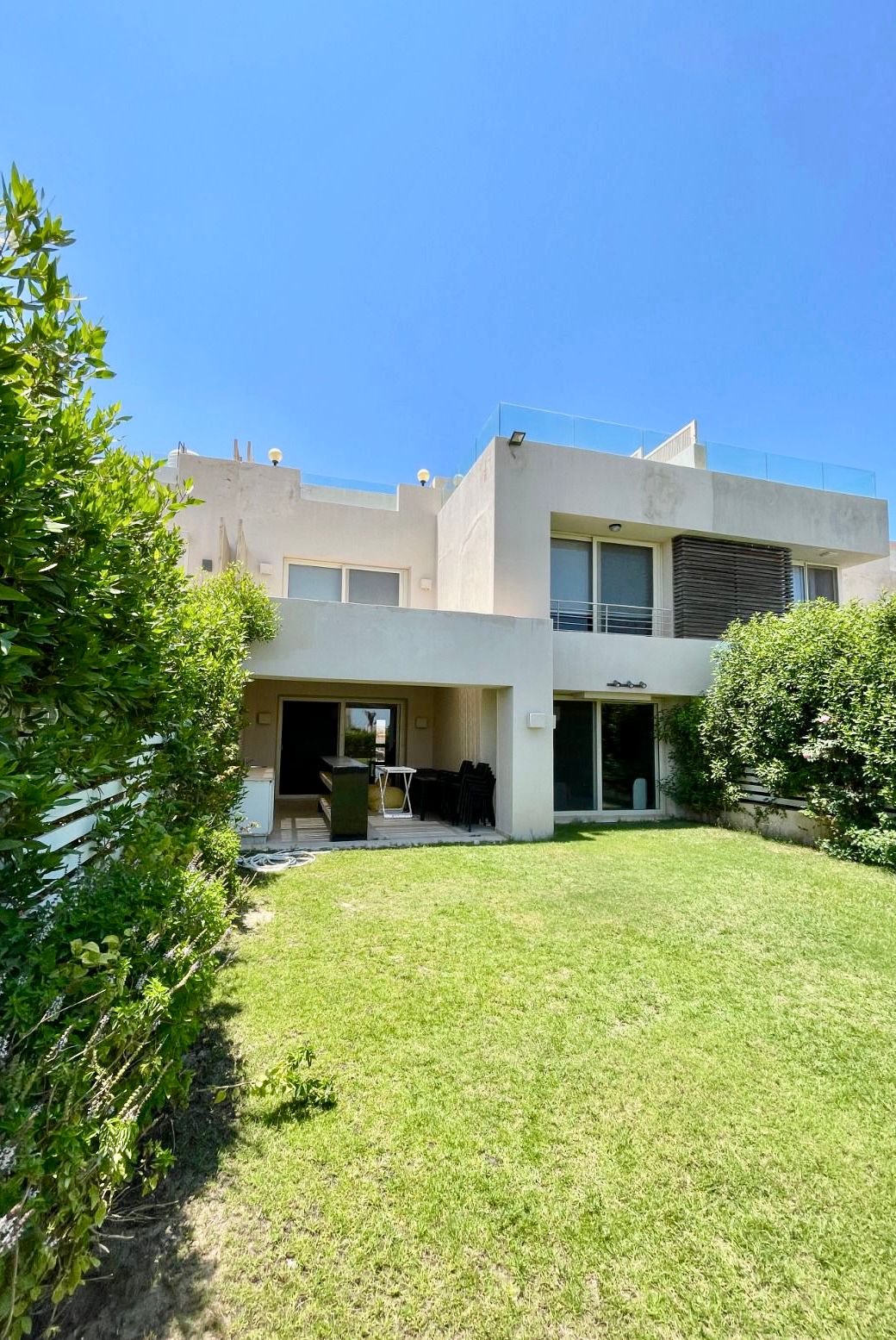 A modern two-story building is surrounded by well-maintained greenery and a spacious lawn. Large glass doors provide an entrance to the interior, while a terrace is visible on the upper level. Bright blue skies enhance the outdoor setting.