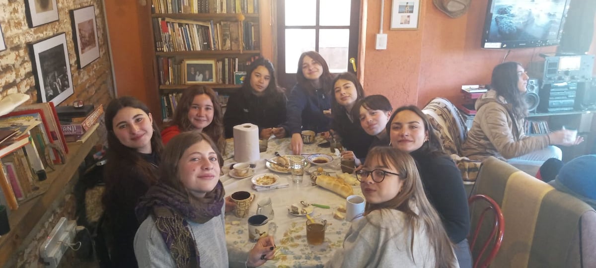 A gathering of individuals is shown around a large table set with various food items and drinks. A bookshelf filled with books lines the wall, and natural light enters through a window, creating a warm and communal atmosphere.