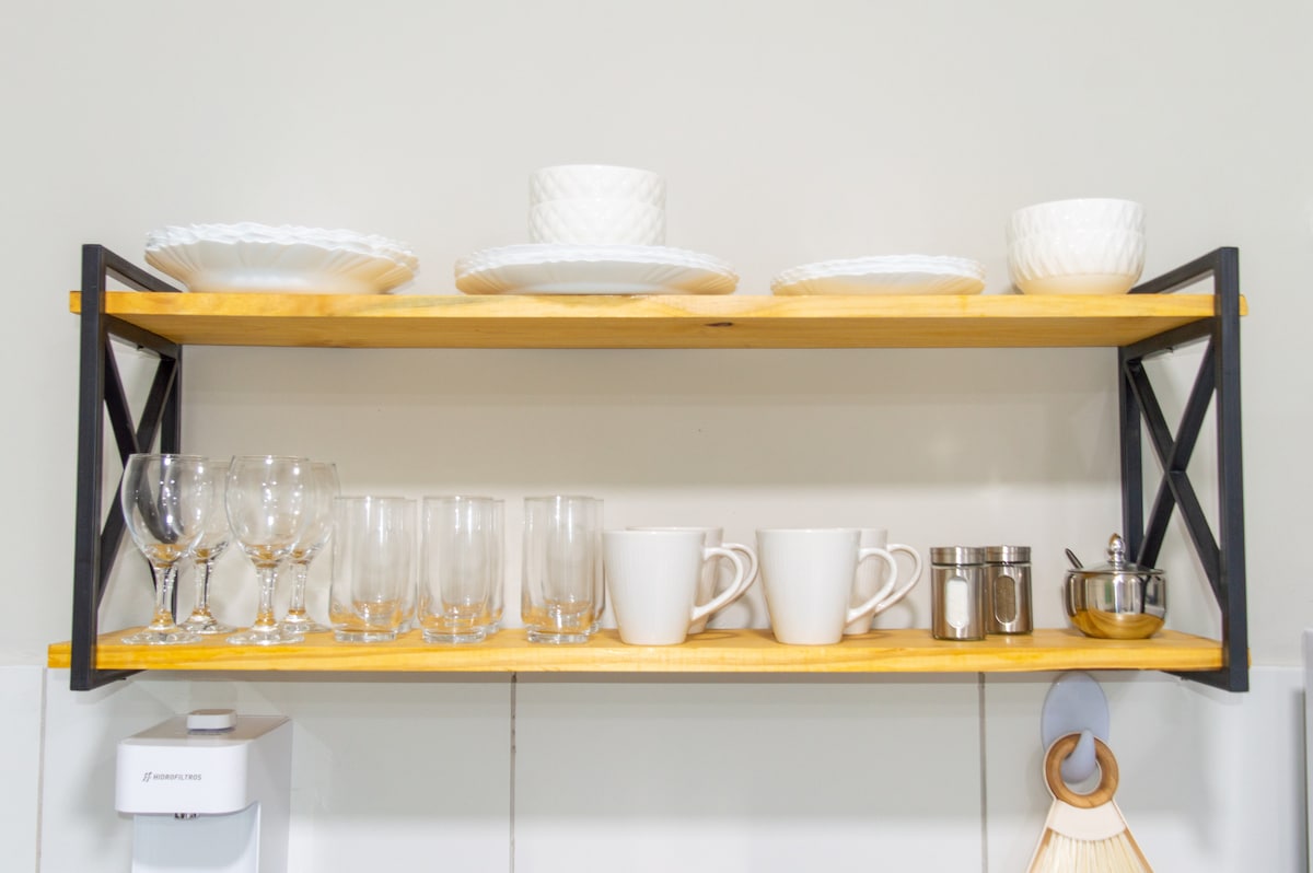 A wooden shelf supported by black metal frames displays an array of white dinnerware, including plates and bowls. Glassware, consisting of both wine glasses and tumblers, aligns with white ceramic mugs and a small sugar bowl. The shelf is neatly organized against a light-colored wall.