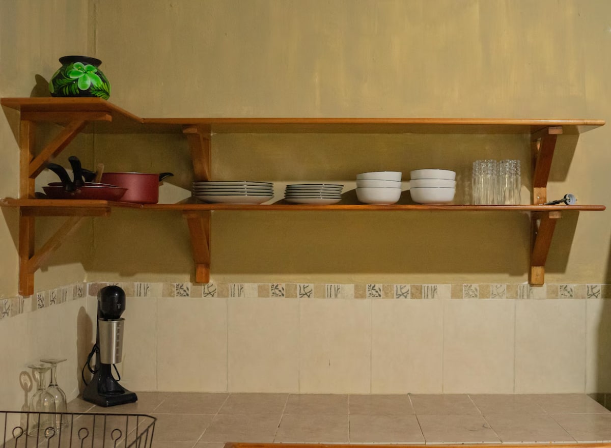 A wooden shelf displays an assortment of kitchenware, including glassware, white bowls, and stacked plates. A red pot can be seen alongside a blender, adding functionality to the space. The kitchen area features tiled walls and a light-colored countertop below the shelves.