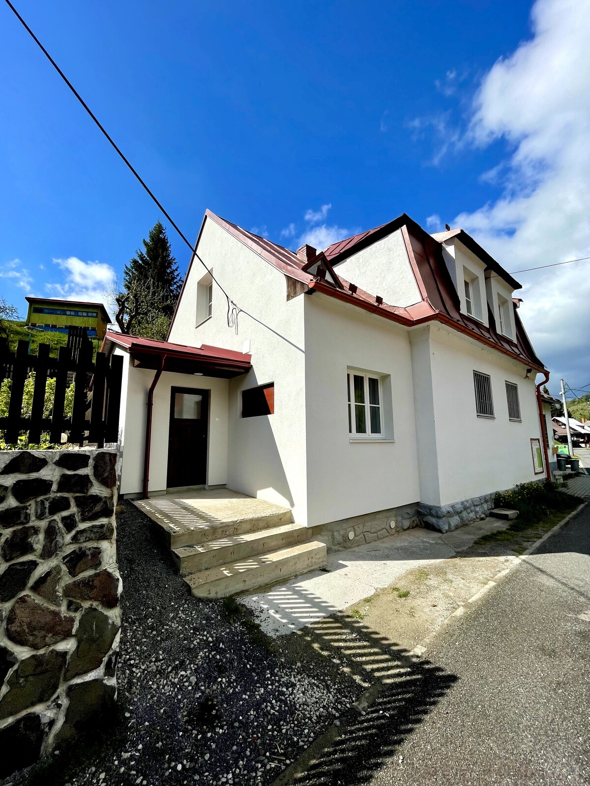 A traditional two-story villa is presented with a white exterior and a distinctive red roof, featuring a welcoming entrance. Natural light illuminates the facade, while the surrounding area includes gravel pathways and a wooden fence. The sky overhead is clear, showcasing a few clouds.