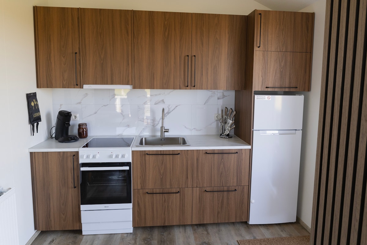A modern kitchen is equipped with wood-finished cabinetry, a stove, and a refrigerator. A sink is located under a window, and utensils are neatly arranged beside it. An electric kettle adds convenience, making this space functional for meal preparation.