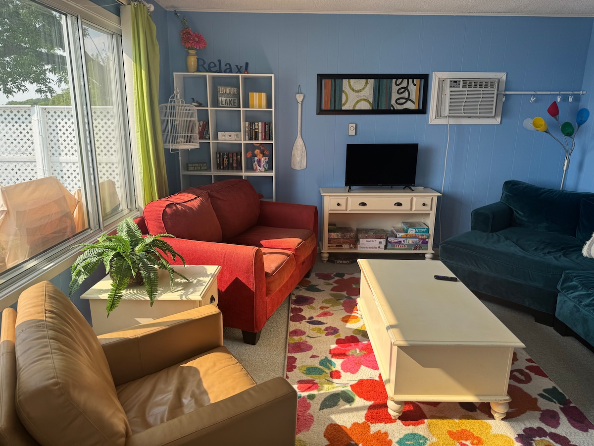 A living area is presented with colorful furnishings, featuring a red armchair and a teal couch. A white coffee table is centered on a vibrant area rug. Natural light illuminates the space through large windows, and a wall-mounted TV is visible on a cream-colored console.
