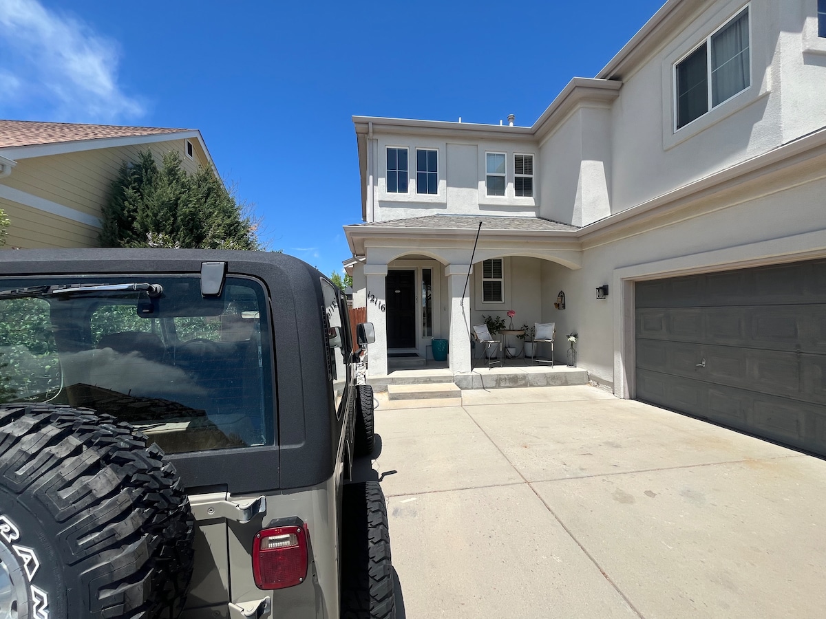 A driveway accommodates up to four cars, with a garage visible on the right. A spacious porch with seating is present at the entrance, surrounded by a welcoming facade of the house. Clear blue skies enhance the setting.