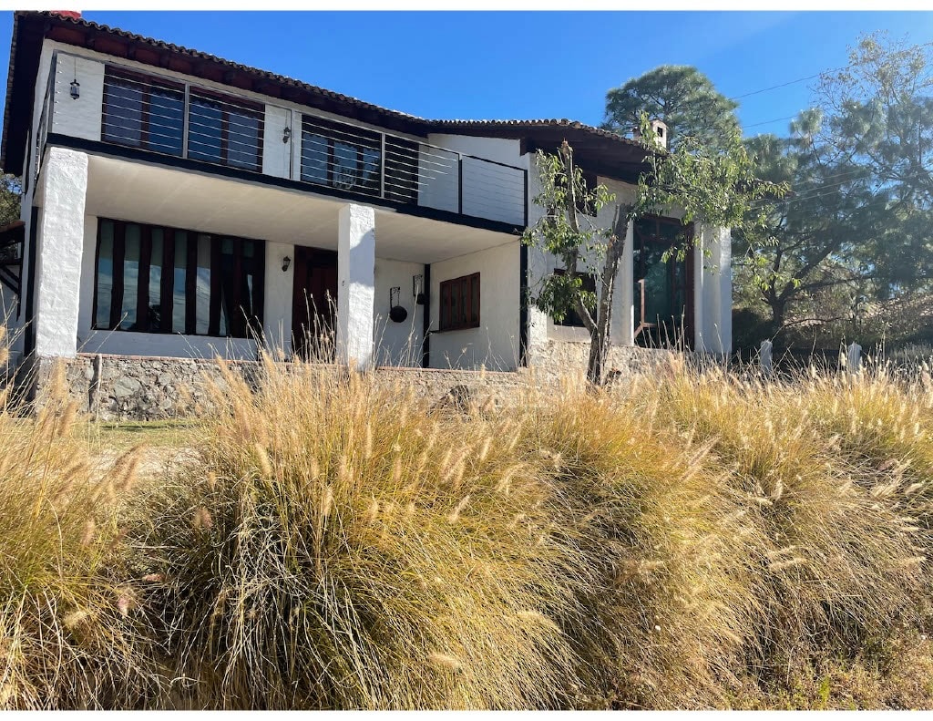 The exterior of a contemporary cabin is captured, featuring a blend of white walls and dark accents. Large glass windows span the upper level, allowing natural light in. Surrounding grass adds texture and warmth, while trees frame the property, emphasizing its connection to nature.