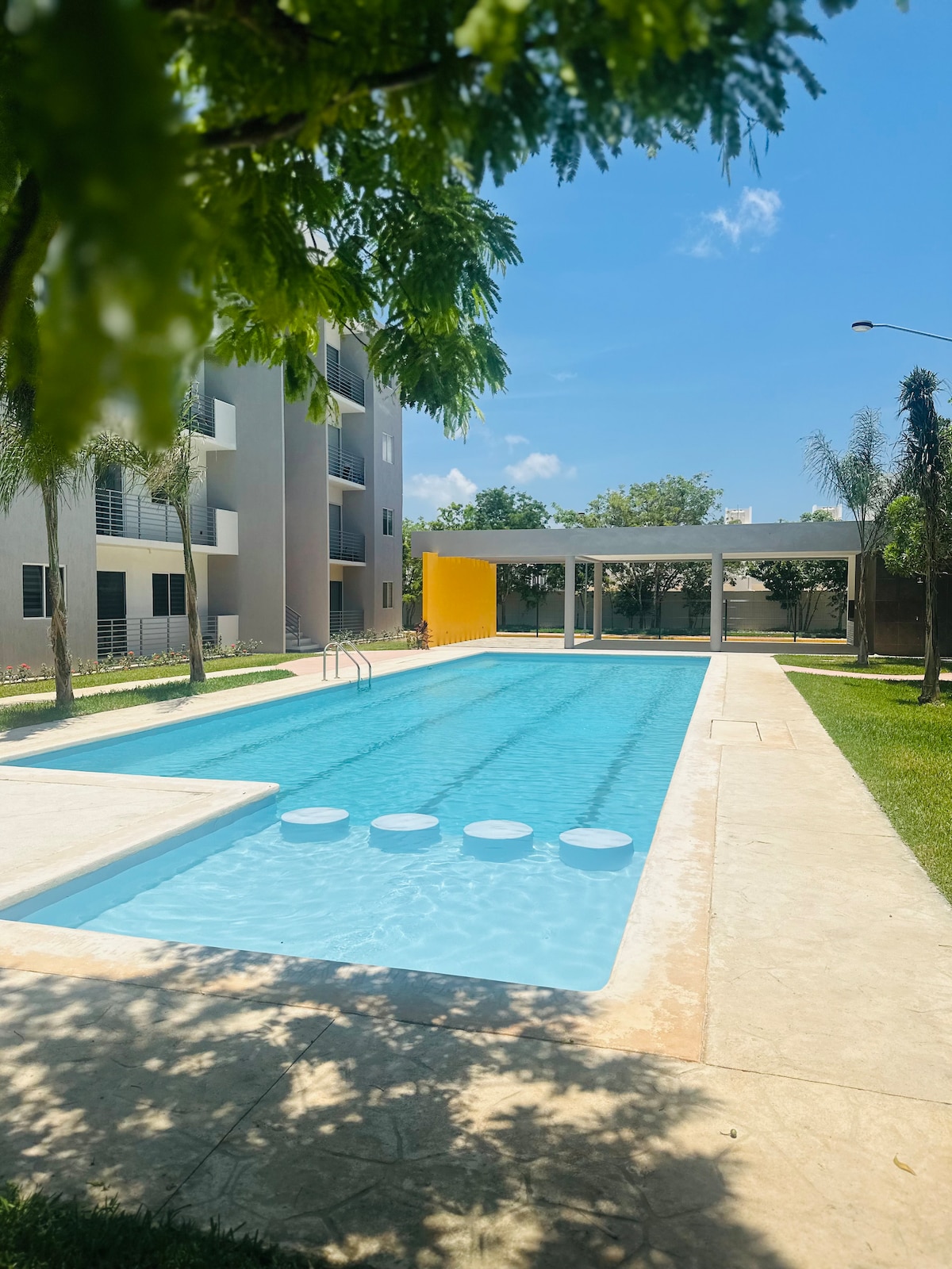 A long, rectangular swimming pool is surrounded by lush greenery and features a sunbathing area. The pool's clear blue water reflects the sky, and white stepping stones are visible in the shallow end. Adjacent to the pool, a shaded area with seating is provided.