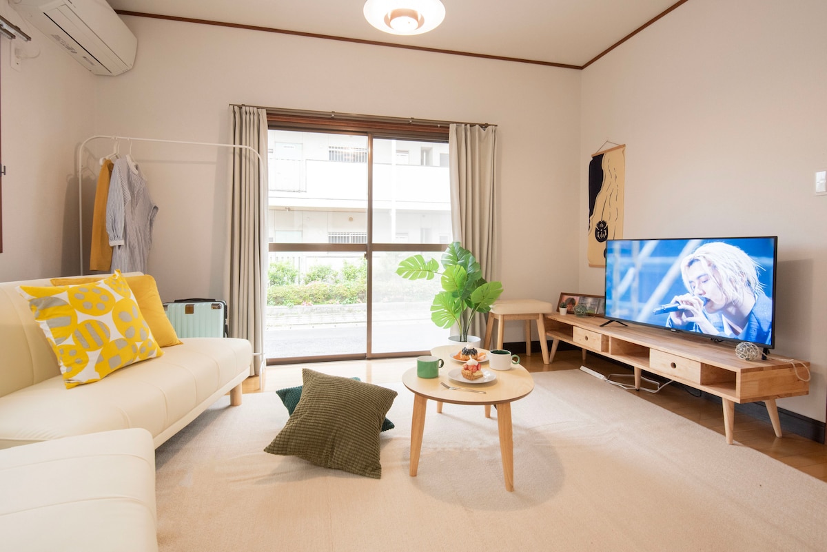 A bright and airy living area is featured with a comfortable sofa and a coffee table. A television displays an artist performing, while a tropical plant adds a touch of greenery. Natural light floods in through large windows adorned with light curtains.