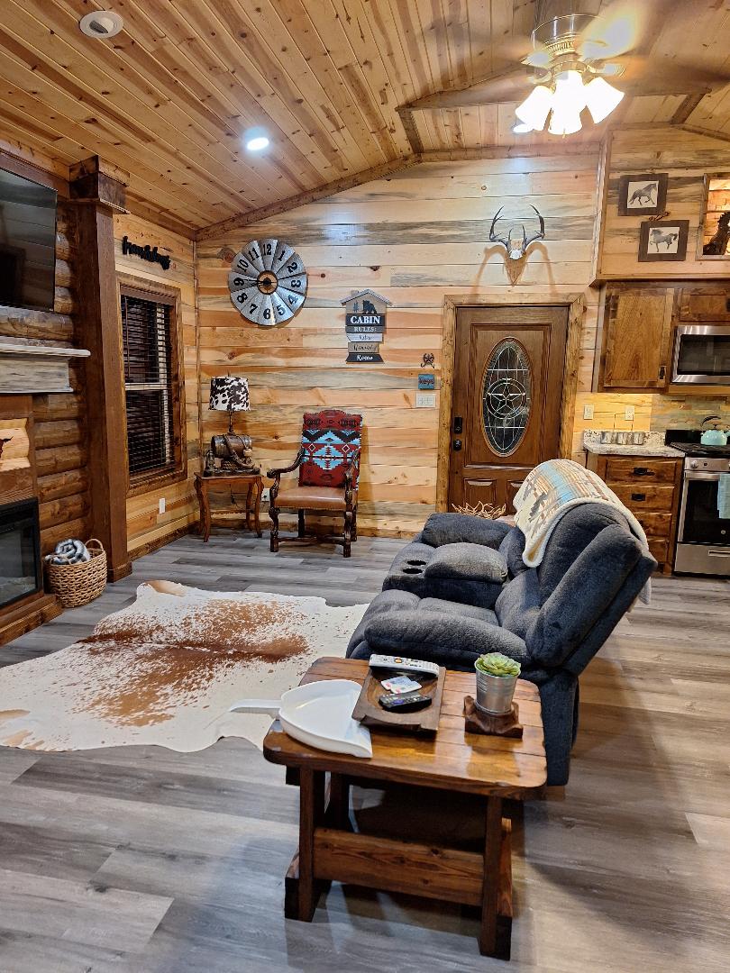 A cozy living area is showcased with wood-paneled walls and a ceiling featuring exposed beams. Comfortable seating is provided by a grey couch, accompanied by rustic decor pieces. A small wooden table holds magazines and a potted plant, while a patterned chair adds charm to the space.
