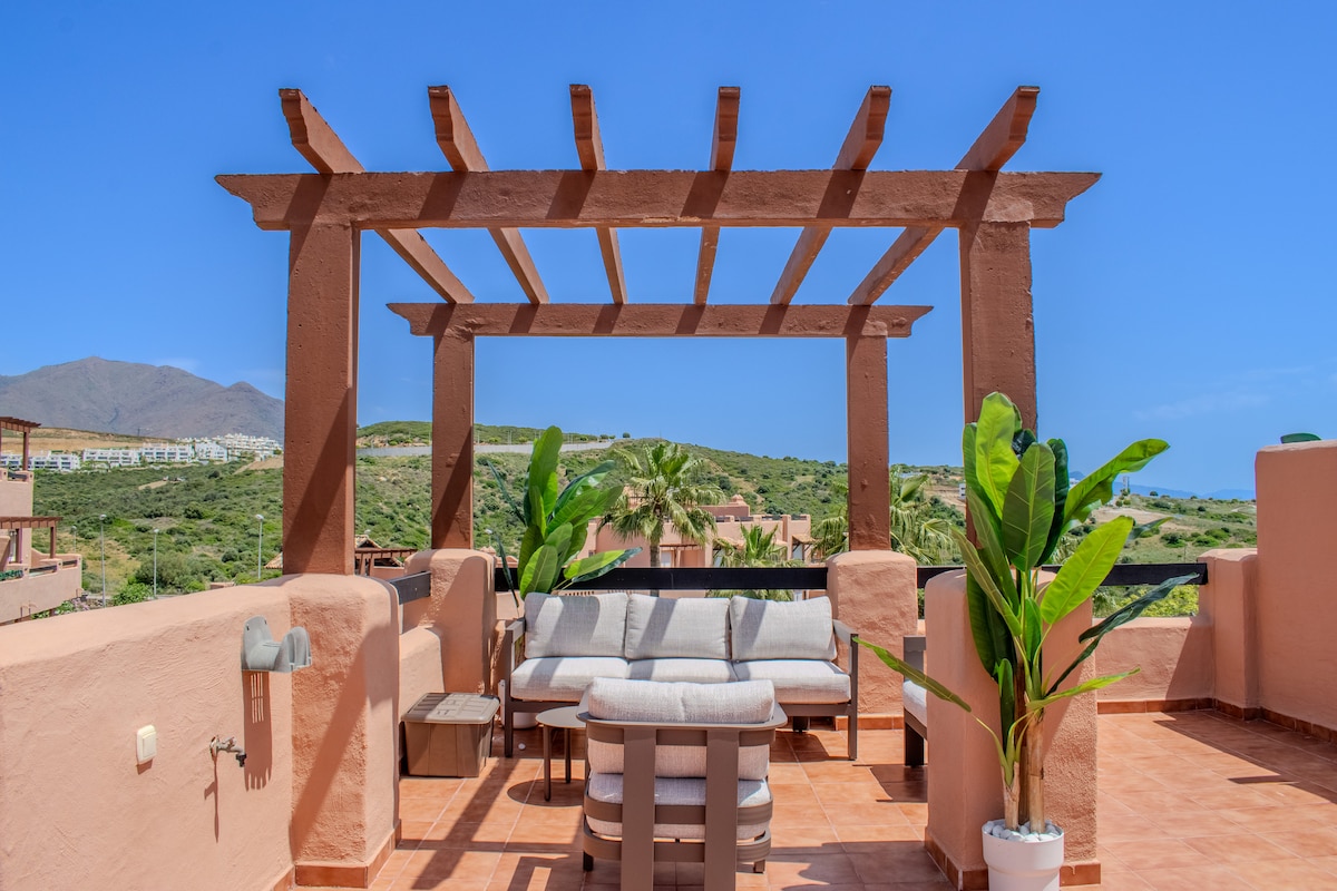 A rooftop terrace features a wooden pergola providing shade over comfortable seating arrangements. Lush green plants are positioned around the area, and panoramic views of surrounding hills and greenery can be seen in the background under a clear blue sky.