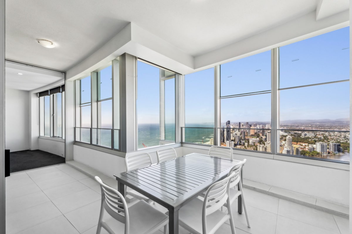 A spacious enclosed balcony offers stunning views of the ocean and surrounding cityscape. A modern dining table with six chairs is positioned to take advantage of the natural light streaming through expansive floor-to-ceiling windows. The tiled floor adds a contemporary touch.