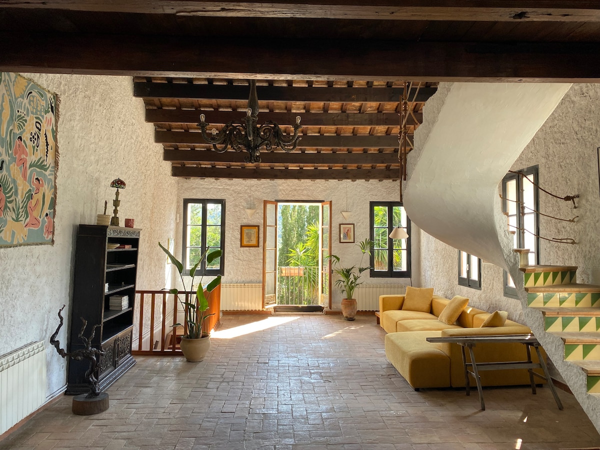 A spacious living area features a combination of rustic and modern elements, highlighted by exposed wooden beams and terracotta flooring. A yellow sofa is positioned toward an inviting doorway that leads to a lush outdoor view. Bookshelves and potted plants add to the welcoming atmosphere.