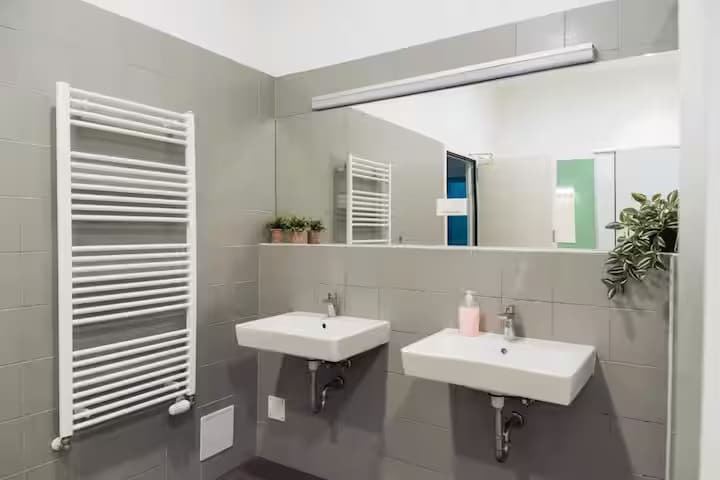 A modern bathroom is equipped with two wall-mounted sinks beneath a wide mirror. A towel warmer extends along one wall, accompanied by small potted plants. Neutral grey tiles create a calm environment, complementing the minimalist design.