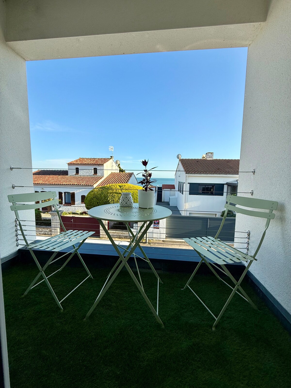 A small balcony features a round table and two folding chairs, surrounded by a vibrant green carpet. In the background, rooftops and a clear blue sky can be seen, creating a pleasant outdoor space for relaxation.