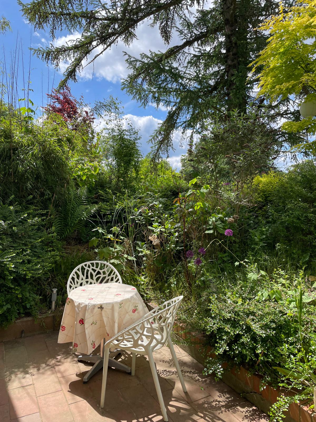 A charming garden space features a small round table covered with a light tablecloth and accompanied by a white chair. Lush greenery surrounds the area, with a variety of plants and flowers creating a peaceful outdoor setting under a bright blue sky.