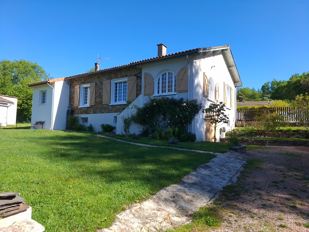 The charming house features a mix of stone and plaster exteriors, framed by green grass and shrubs. A pathway of stone leads to the entrance, and several windows with wooden shutters create a welcoming facade. Lush greenery surrounds the property, enhancing its serene setting.