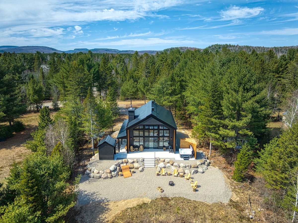 An aerial view captures the modern chalet nestled among lush greenery, highlighting a spacious outdoor area. Comfortable seating is arranged around a fire pit, while a luxurious hot tub is positioned nearby, inviting relaxation in a serene natural landscape.