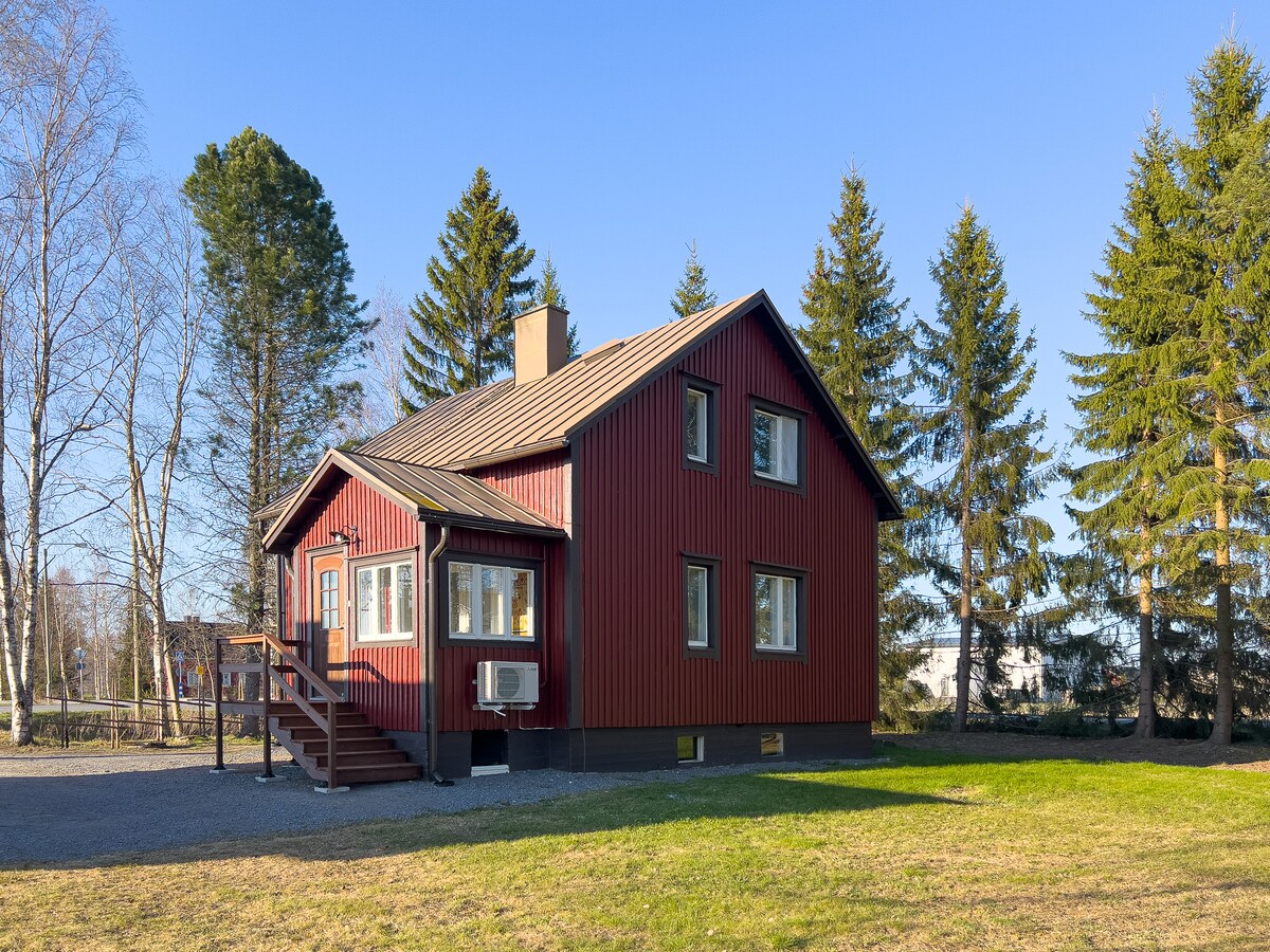The exterior of a traditional red wooden house is displayed, featuring a sloped roof and multiple windows. The house is surrounded by green grass, tall pine trees, and a gravel pathway leading to the entrance. Clear blue skies create a serene backdrop.