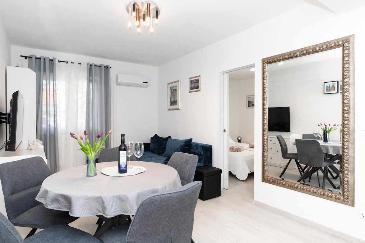 A dining area features a round table set with two glasses and a wine bottle. Surrounding the table are four upholstered chairs. A comfortable blue sofa is positioned nearby, and a large mirror reflects the bright lighting and decor in the room.