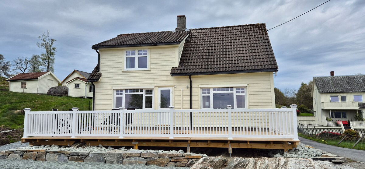 A charming two-story house features a light yellow exterior and a dark tiled roof. A spacious deck with white railings offers an outdoor seating area. Large windows allow natural light to fill the interior, and a well-maintained landscape surrounds the property.