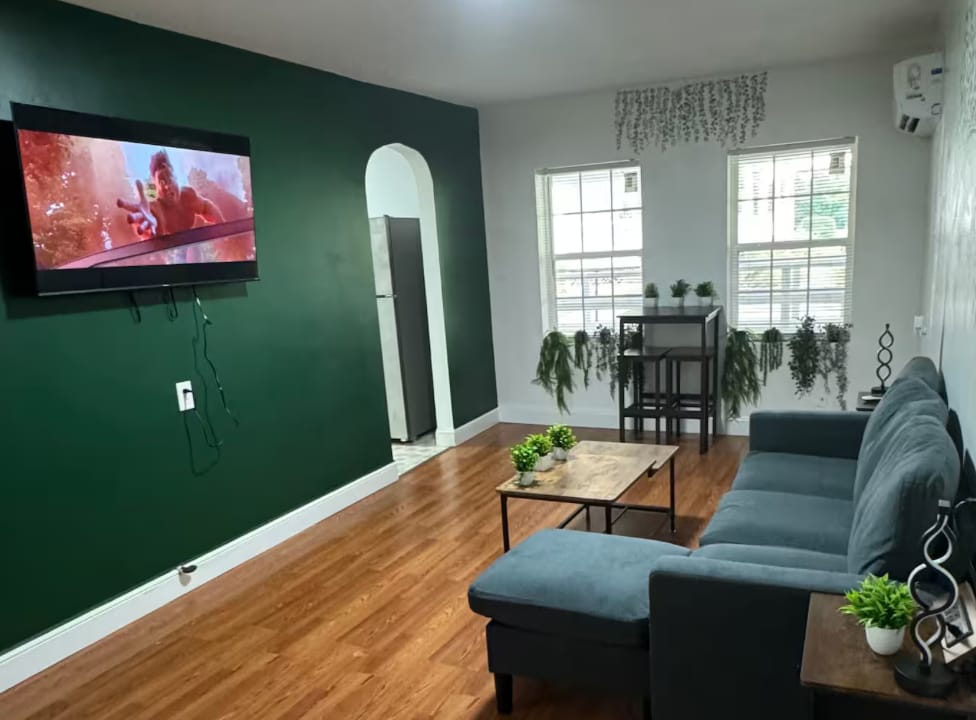 A cozy living room is showcased, featuring a dark green accent wall and large windows with greenery. A comfortable couch faces a wall-mounted television. A small dining table for two sits adjacent, surrounded by indoor plants, creating a serene and inviting environment.