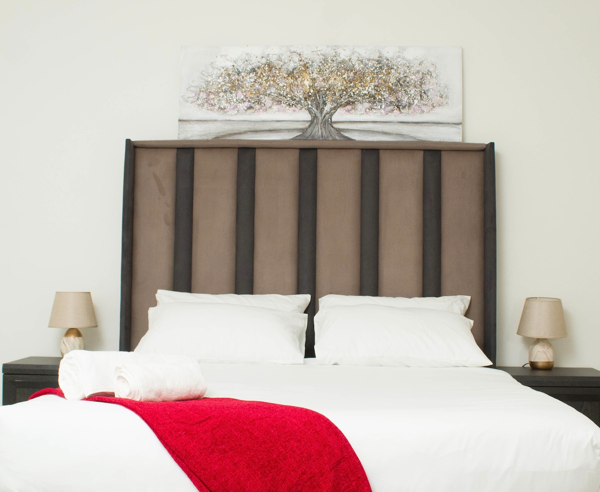 A spacious bed is positioned against a light-colored wall, topped with decorative pillows. An artistic depiction of a tree hangs above the headboard. Two bedside lamps are placed on each side, and a rolled towel is neatly arranged on the bed, accompanied by a vibrant red blanket.