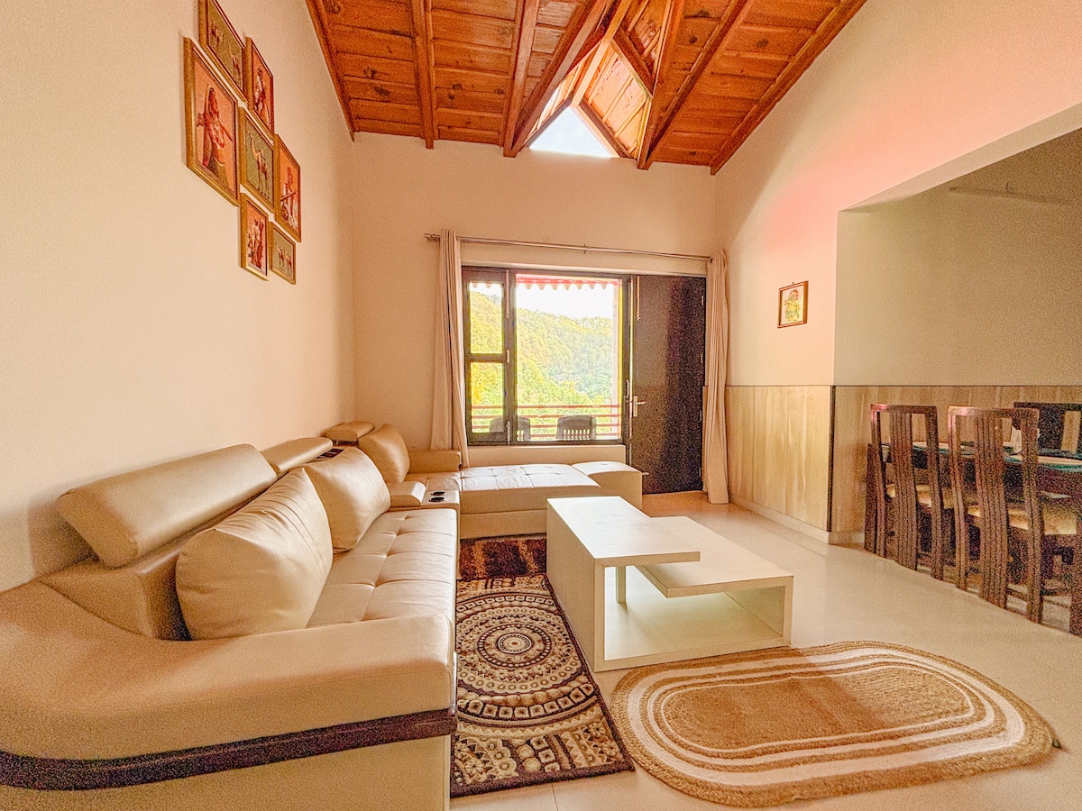 A light-filled living area features a comfortable cream-colored sofa and a low coffee table on a patterned rug. Large windows showcase scenic views of the surrounding hills. A dining area with wooden chairs is visible in the background, complemented by a wooden ceiling.